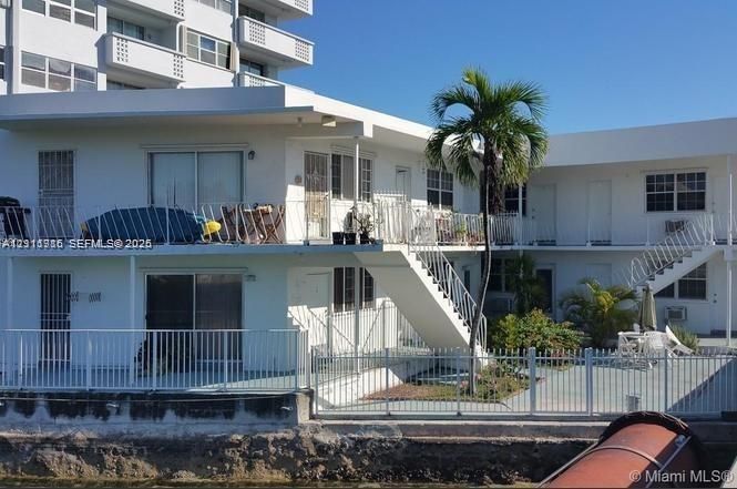 Waterfront building exterior featuring multi-level open-air balconies, metal railings, sliding glass doors, and a palm-lined concrete patio
