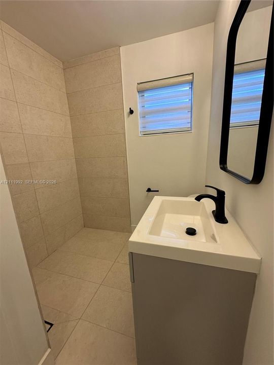 Walk-in shower with floor-to-ceiling neutral tile, modern vanity with a rectangular sink, matte black faucet, wall-mounted mirror with a black frame, and window with integrated blinds