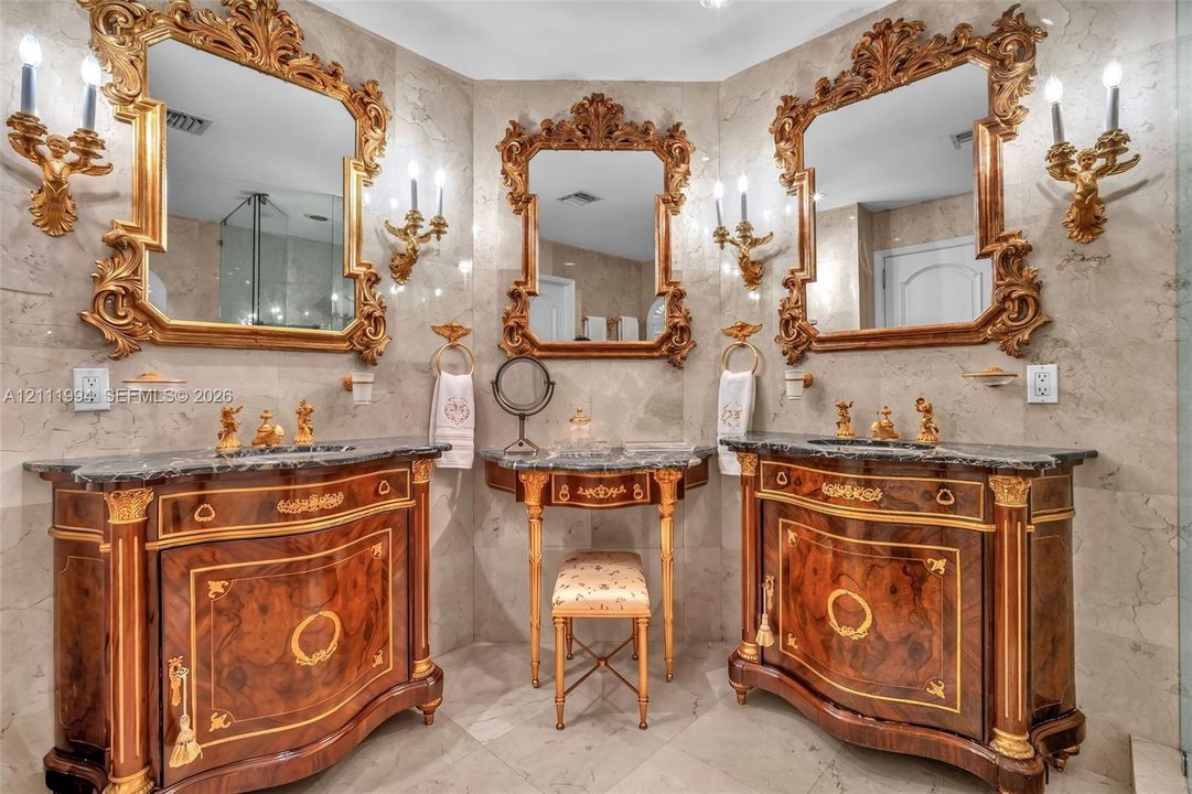 Custom dual vanities with dark stone countertops, ornate gold-leaf framed mirrors, integrated central makeup vanity, gold-tone wall-mounted candle-style sconces, and neutral tile flooring