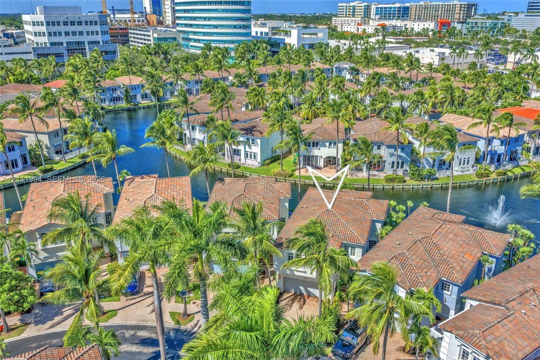 Aerial perspective of a multi-story residence featuring a terracotta-tiled roof, concrete exterior, attached two-car garage, and waterfront positioning along a canal with manicured landscaping and mature palm trees