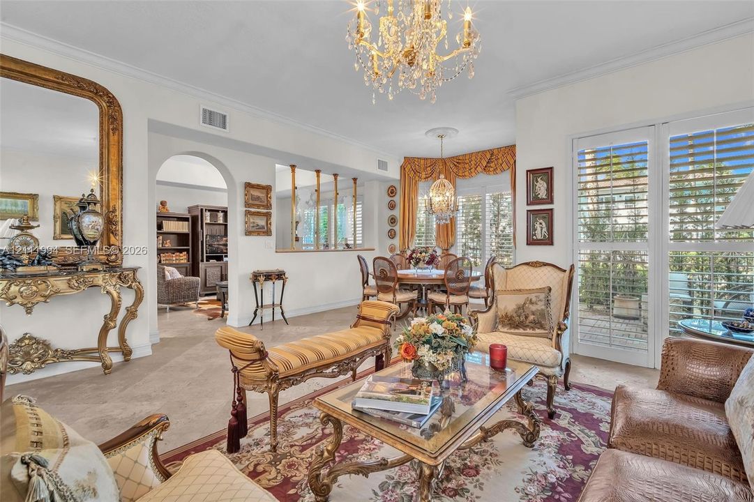 Interior space featuring architectural archways, a tiered display cutout with columns, crown molding, multi-pane glass doors with plantation shutters, and ornate crystal chandeliers