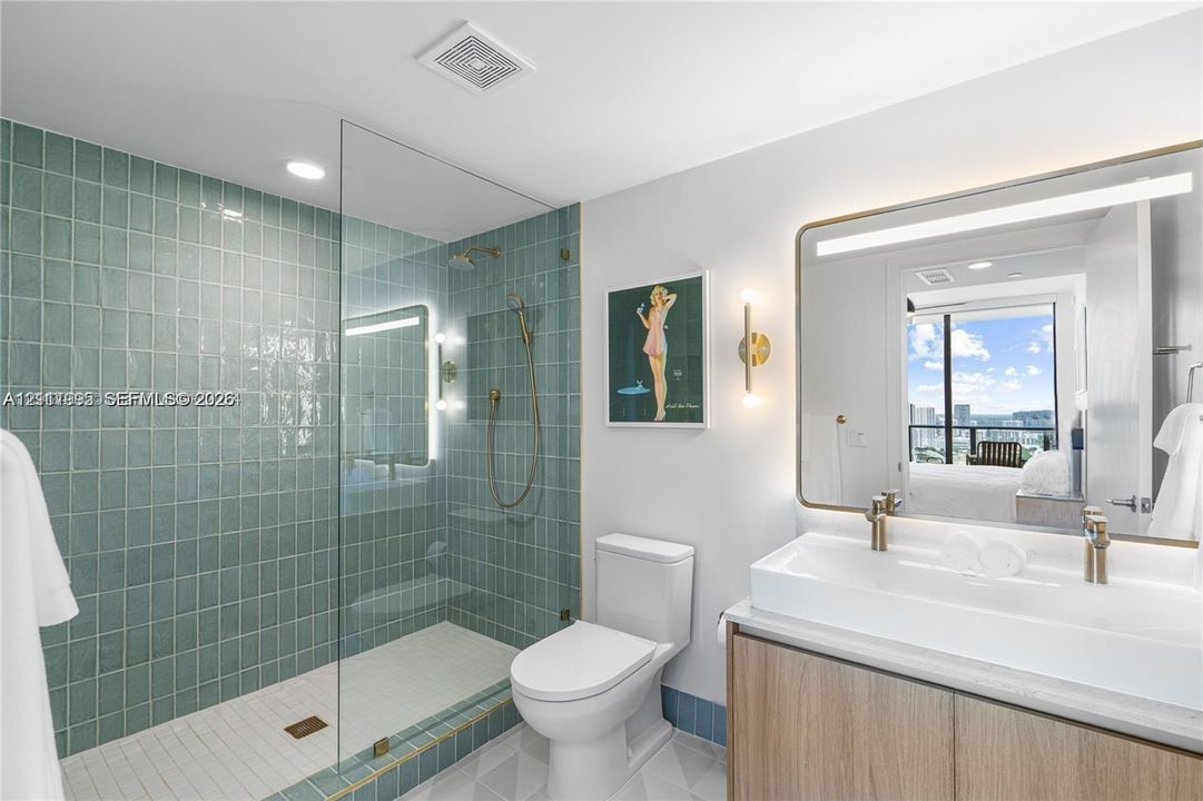 Bathroom featuring a walk-in shower with seafoam-colored tiled walls, glass enclosure, wood-finish vanity with a dual-faucet trough sink, and an expansive backlit mirror