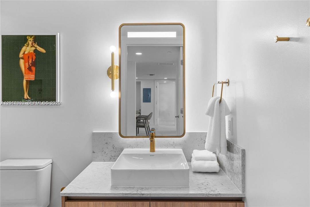 Rectangular vessel sink with a brass faucet, stone-finish countertop, backlit mirror with brass frame, and wall-mounted brass light fixture