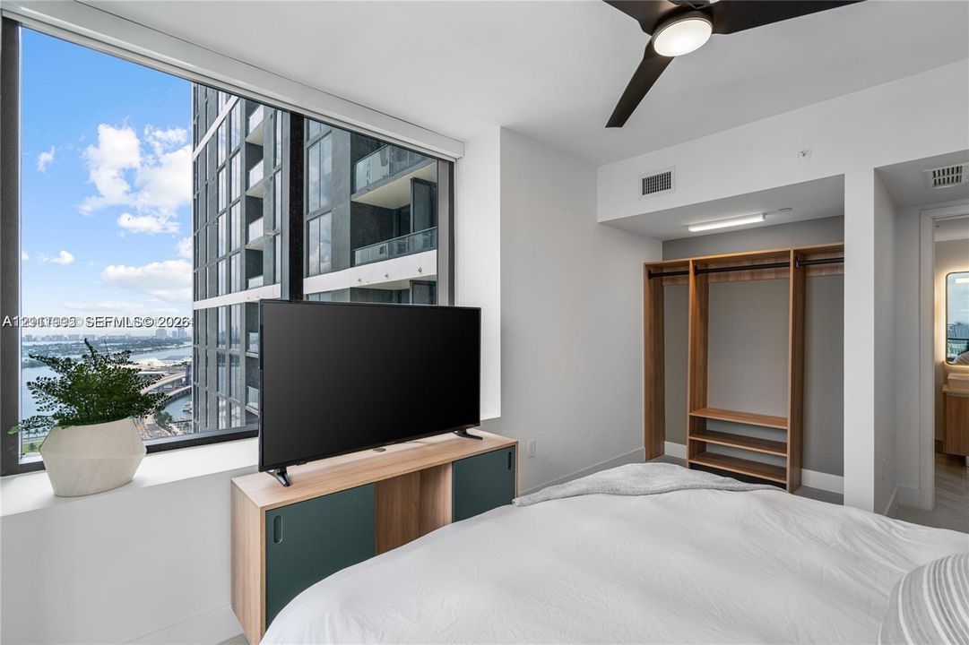 Floor-to-ceiling windows, built-in wooden wardrobe shelving, and a ceiling-mounted light fixture with a fan