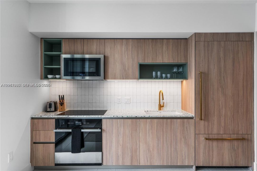 Kitchen cabinetry with wood-finish flat-panel doors, speckled stone countertop, white grid-pattern tile backsplash, integrated stainless steel appliances, and gold-tone hardware