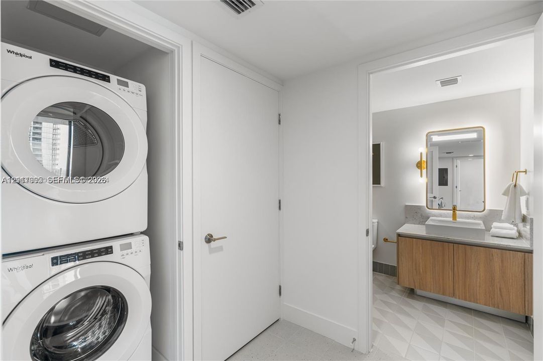 Stacked laundry center featuring high-efficiency white appliances, floating wood-finish vanity with a rectangular vessel sink, stone-surface countertop, and illuminated wall mirror