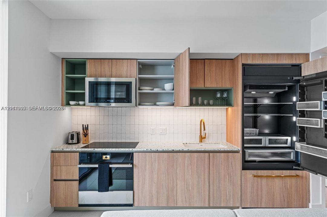 Kitchen featuring wood-finish cabinetry with open accent shelving, integrated refrigerator, stone-composite countertop, white square-tile backsplash, and brass-tone faucet