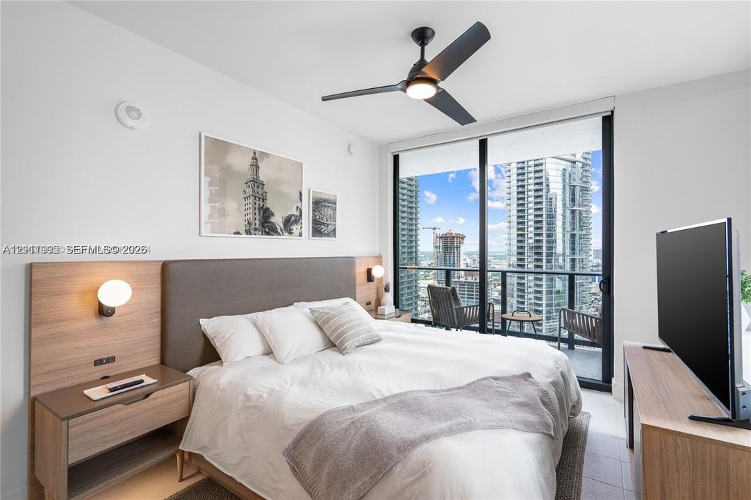 Floor-to-ceiling windows with balcony access, wood-finish built-in headboard with integrated lighting, and a modern ceiling fan