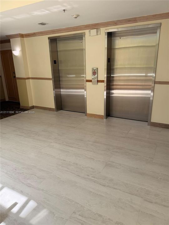 Common area elevator lobby featuring dual stainless steel elevator doors, large-format tile flooring, wood chair rail molding, and wall-mounted sconce lighting