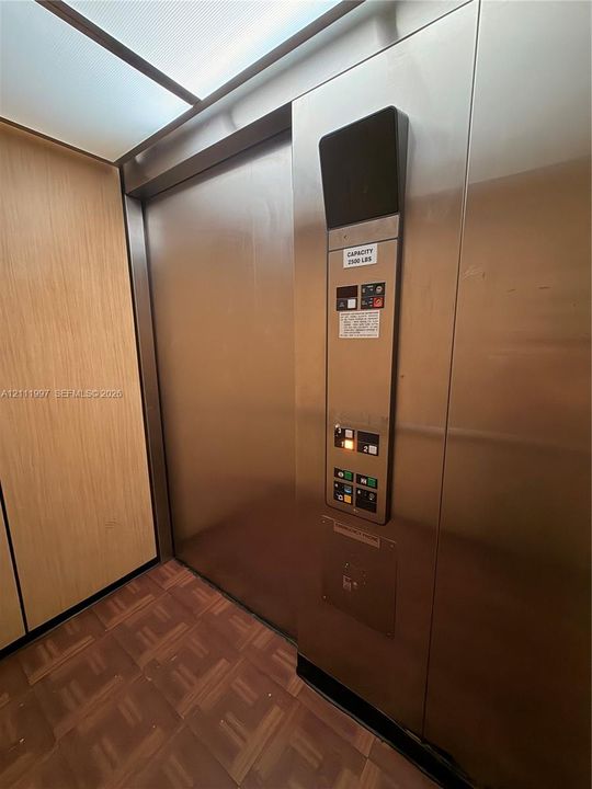 Elevator interior featuring brushed stainless steel panels, wood-finish wall paneling, parquet flooring, and an integrated ceiling lighting panel
