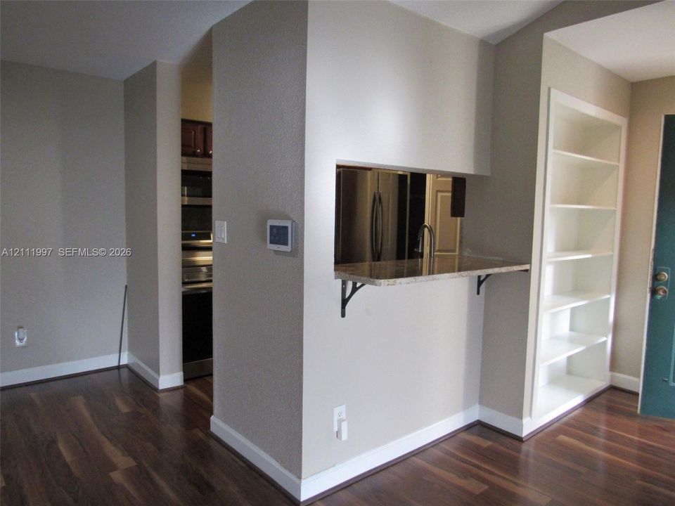 Built-in shelving unit, wood-finish flooring, pass-through opening with a stone countertop, and stainless steel kitchen appliances
