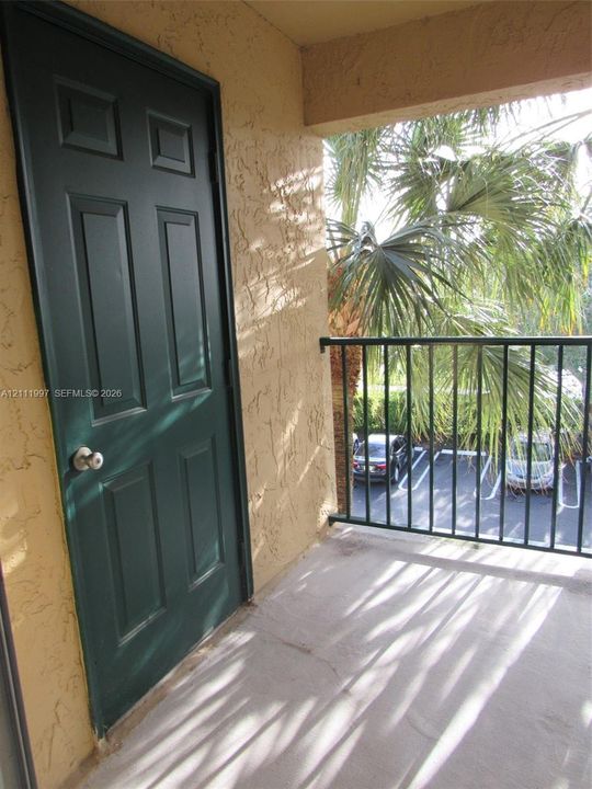 Exterior balcony featuring a smooth concrete floor, textured stucco walls, a six-panel door with a metallic hardware finish, and a black metal railing