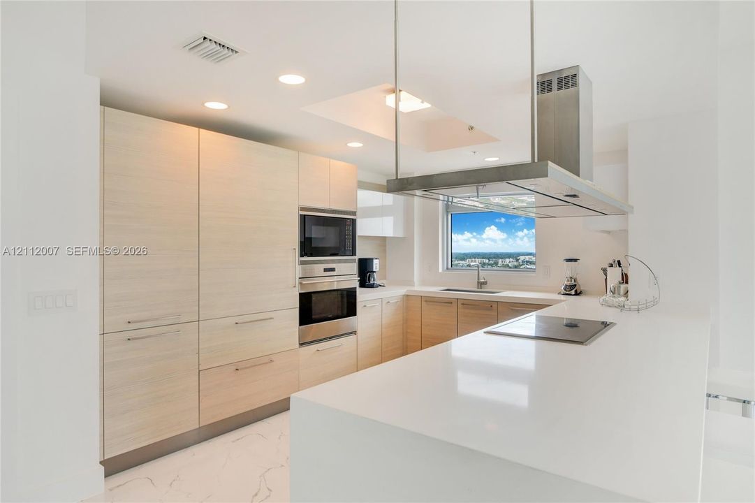 Kitchen featuring flat-panel wood-finish cabinetry, white solid-surface countertops, a stainless steel ceiling-mounted vent hood, built-in stainless steel double ovens, and large-format flooring with a veined pattern
