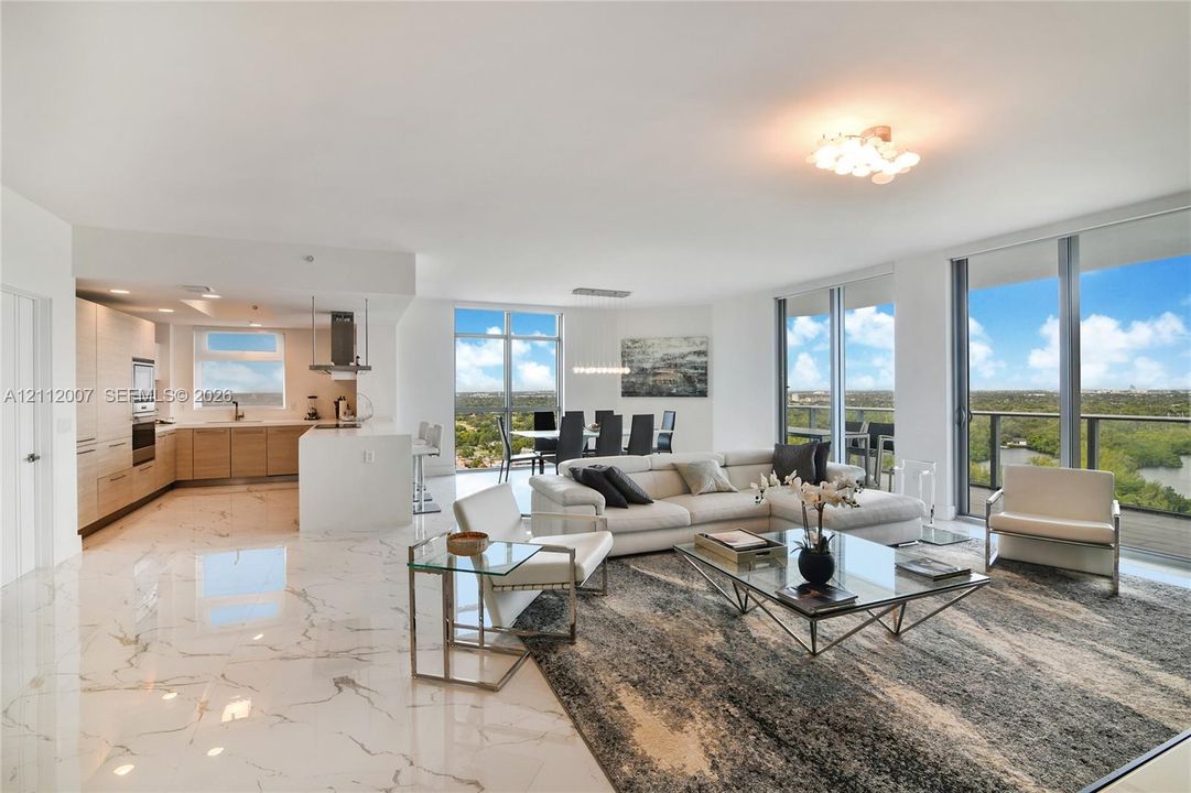Open-concept interior featuring large-format polished stone flooring, floor-to-ceiling glass sliding doors, built-in light-tone cabinetry, a central kitchen island, and a balcony