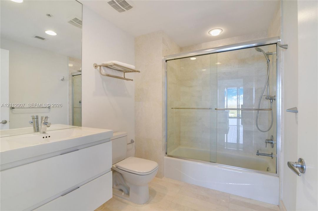 Bathroom featuring a modern floating vanity with a white countertop, a shower and tub combination with a glass enclosure, polished chrome fixtures, and neutral stone tile flooring