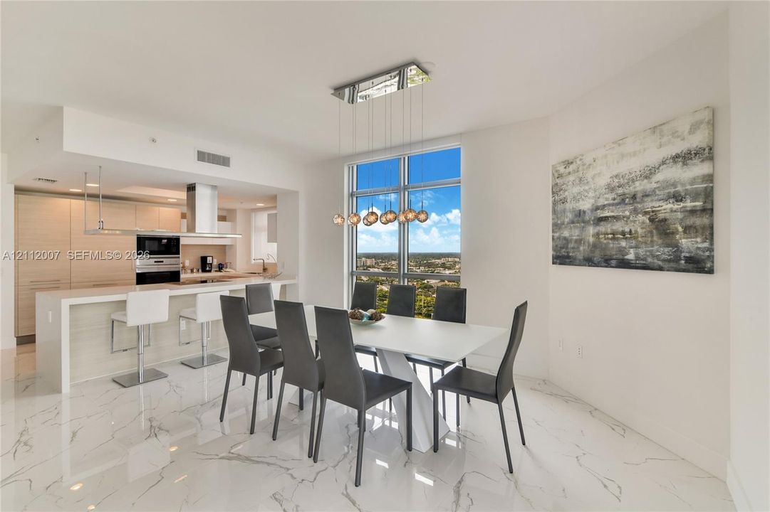 Open-concept interior featuring light-toned cabinetry, stainless steel built-in appliances, floor-to-ceiling windows, polished stone-finish tile flooring, and a multi-bulb linear pendant light fixture