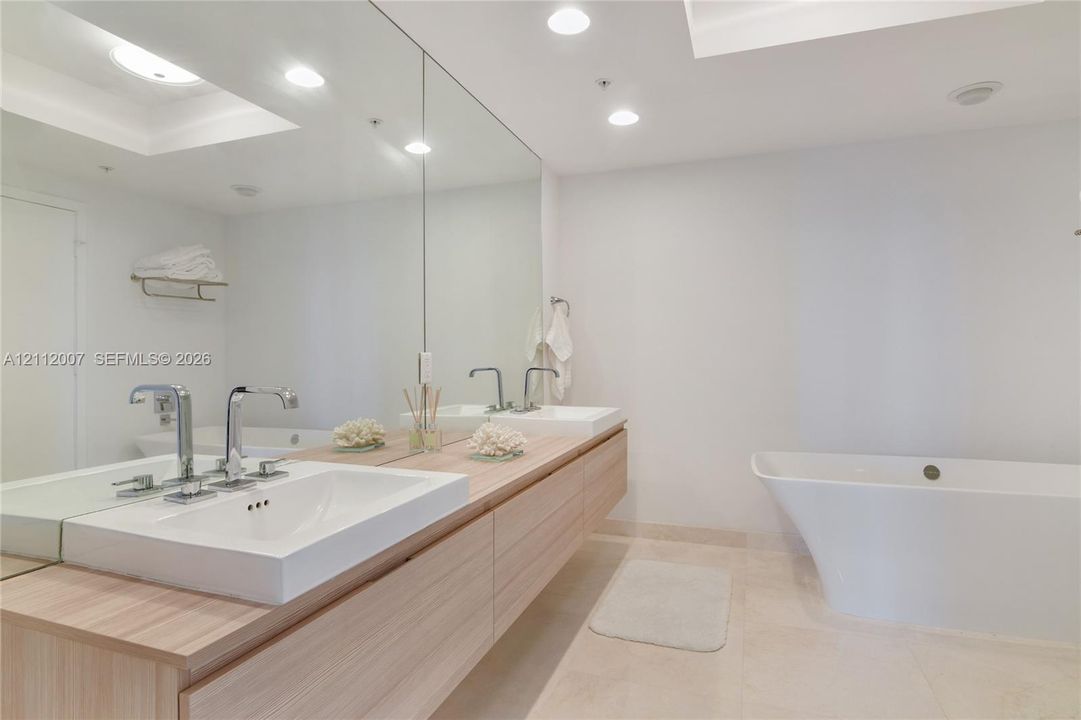 Floating light wood-finish vanity with dual rectangular sinks, polished chrome widespread faucets, full-width wall mirror, and freestanding soaking tub