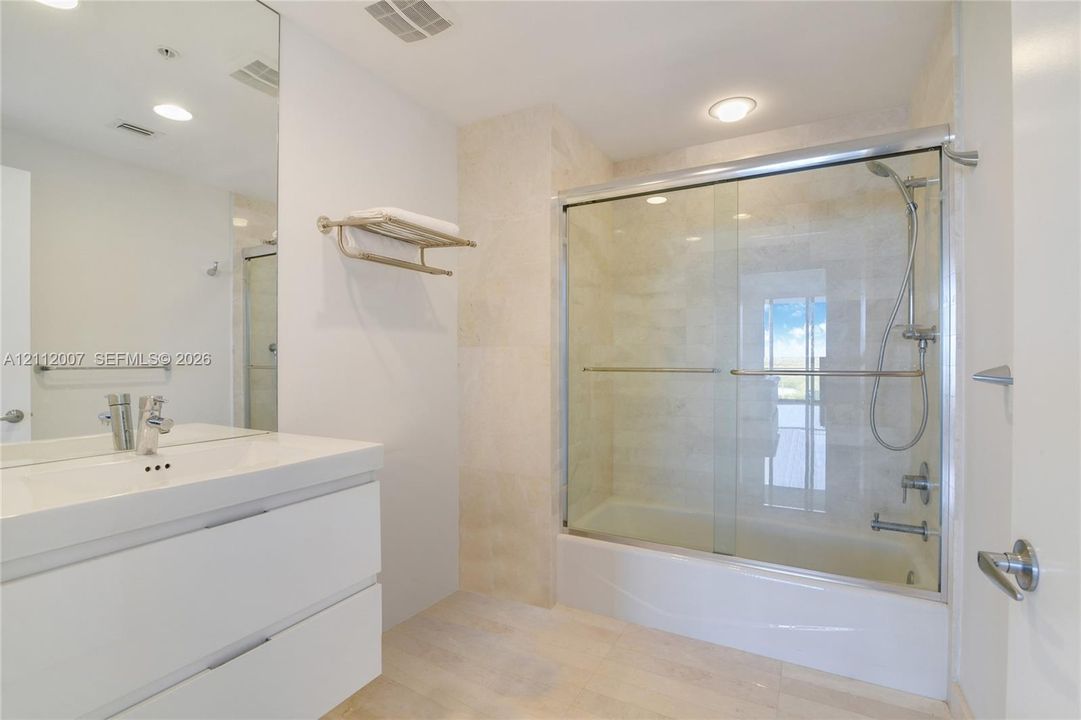Modern vanity with white cabinetry, integrated sink, and polished chrome faucet, featuring a wall-spanning mirror, glass-enclosed shower with a combination tub, and large-format tile surround