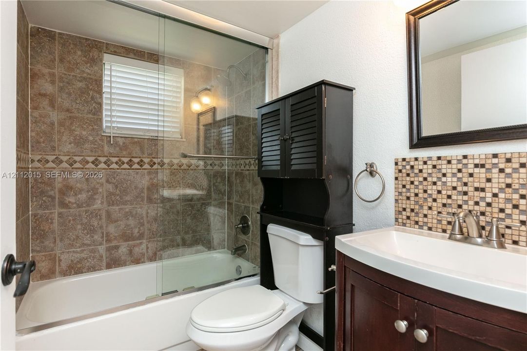 Bathroom featuring a shower-tub combination with glass enclosure, tiled surround with decorative accent border, mosaic tile backsplash, dark wood-finish vanity, and wall-mounted mirror