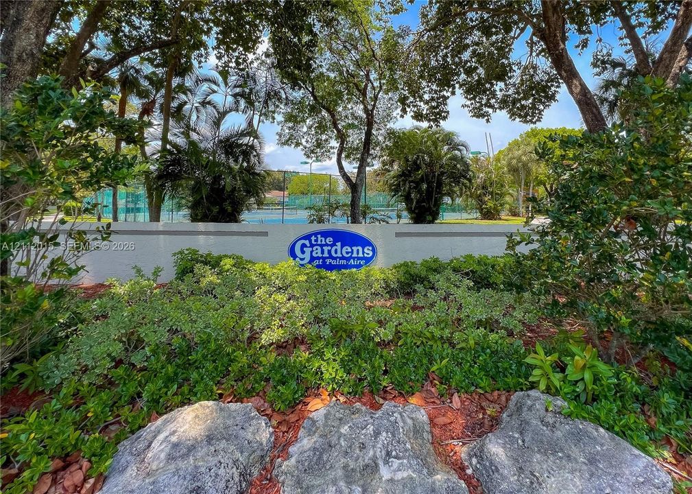 Landscaped entrance featuring mature shade trees, stone boulders, shrubbery, and signage for The Gardens at Palm-Aire