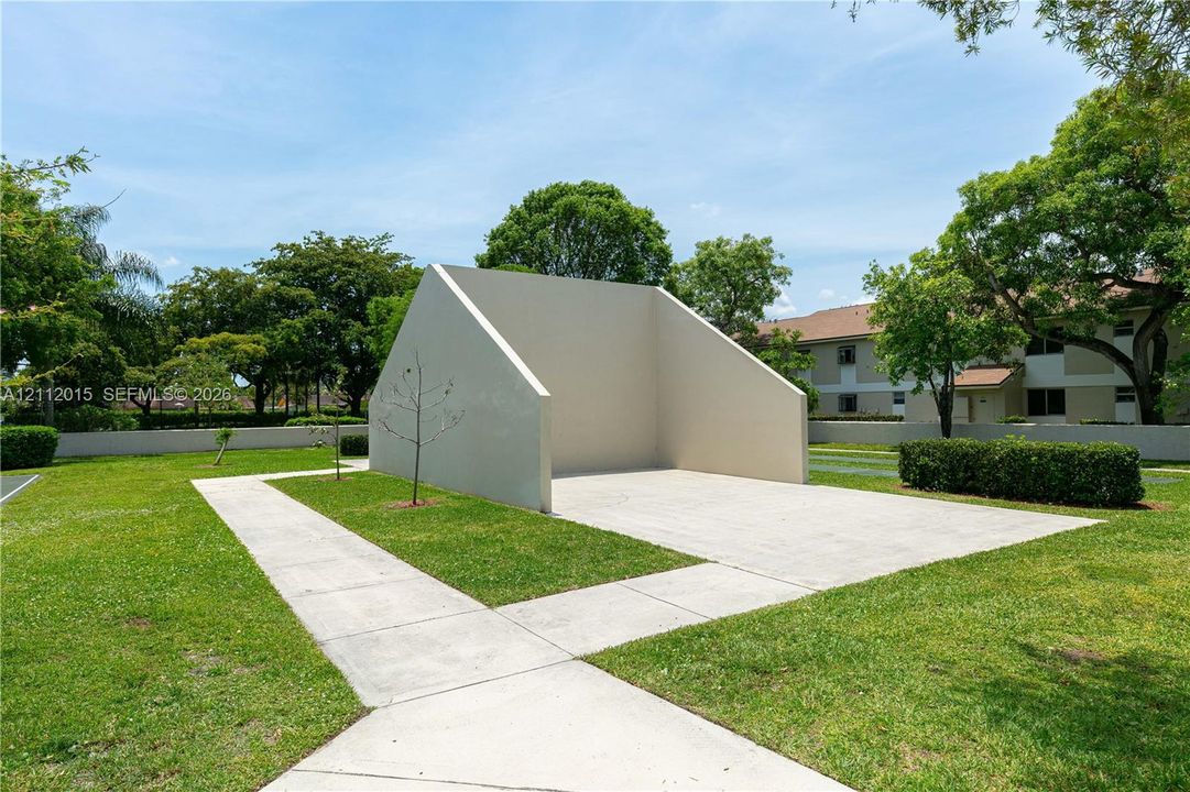 Handball court featuring a concrete surface and a solid stucco-finish backboard situated within a lawn area with paved pathways