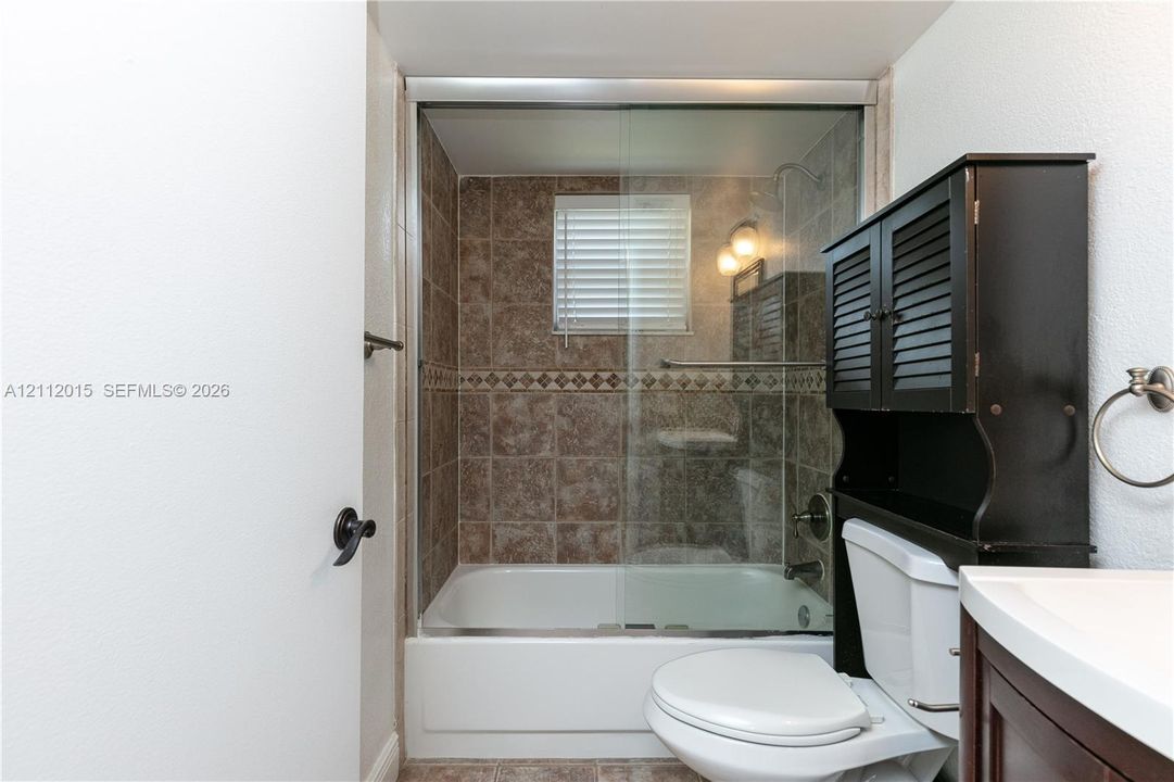 Bathtub with glass sliding enclosure, tile wall surround with decorative accent inlay, built-in over-the-toilet storage cabinet, and wall-mounted towel ring