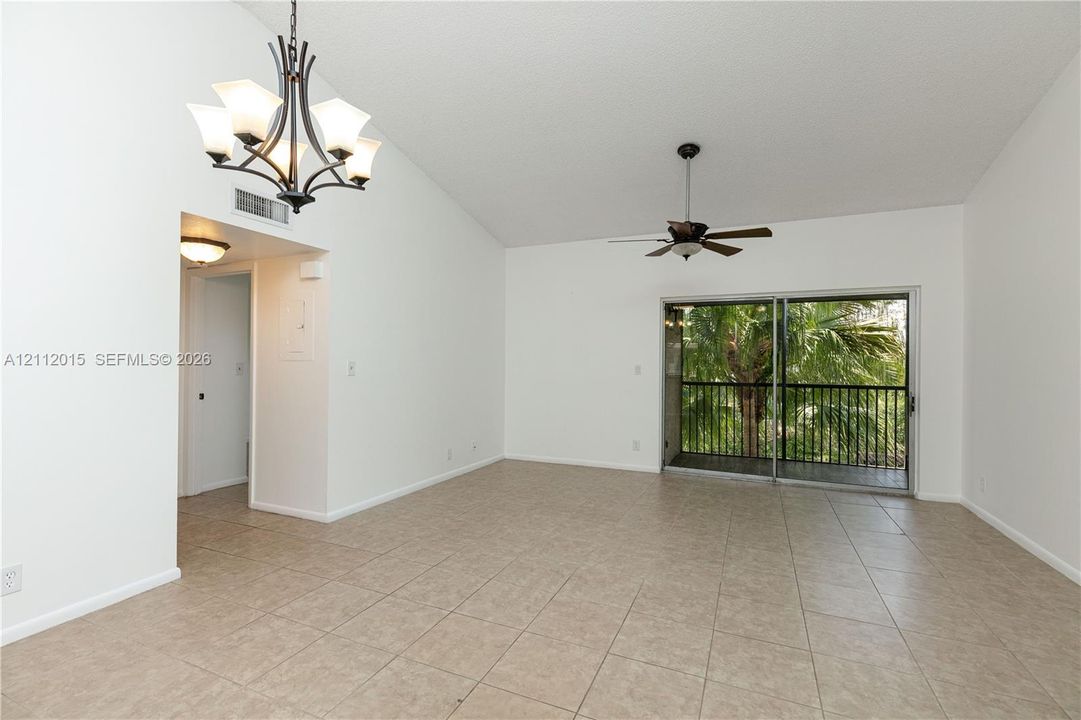 Vaulted ceilings, neutral tile flooring, multi-light iron chandelier, ceiling fan, and sliding glass door leading to a railed balcony