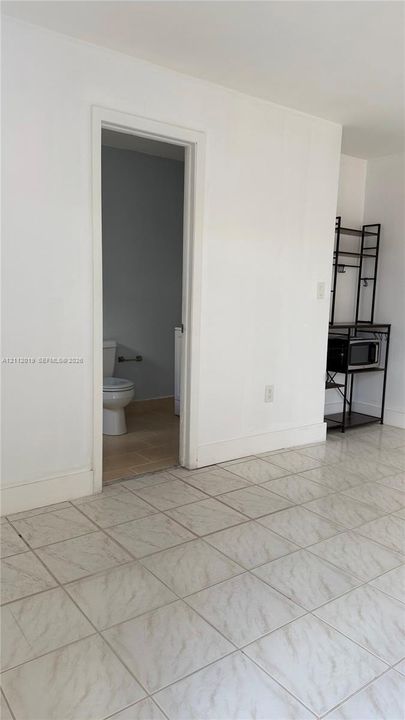 Interior space featuring large-format neutral tile flooring throughout and a doorway revealing a restroom with a toilet and tile flooring