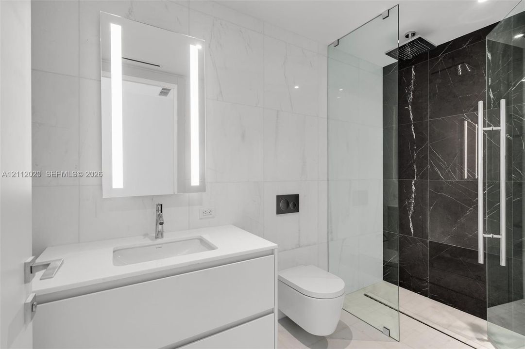 Wall-mounted vanity with a white countertop and rectangular undermount sink, illuminated vertical mirror, wall-hung toilet with a dual-flush actuator, and walk-in shower featuring a glass enclosure and dark tile surround