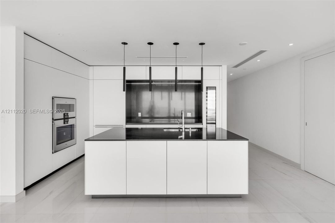 Minimalist kitchen featuring flat-panel cabinetry, a central island with a contrasting stone countertop, built-in stainless steel double ovens, a metal backsplash, and suspended cylindrical pendant lighting