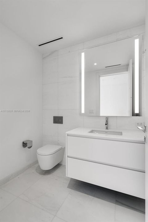 Modern floating vanity featuring a smooth-surface countertop, wall-mounted commode, large integrated LED-lit mirror, and large-format wall and floor tiles