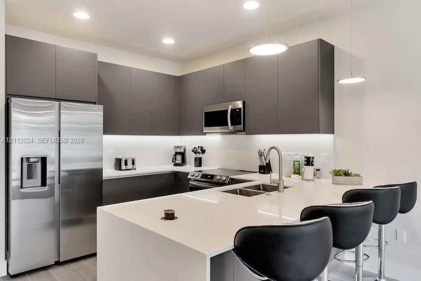 Modern kitchen featuring matte grey flat-panel cabinetry, white solid-surface countertops, stainless steel appliances, recessed ceiling lighting, and a kitchen island with an integrated stainless steel dual-basin sink