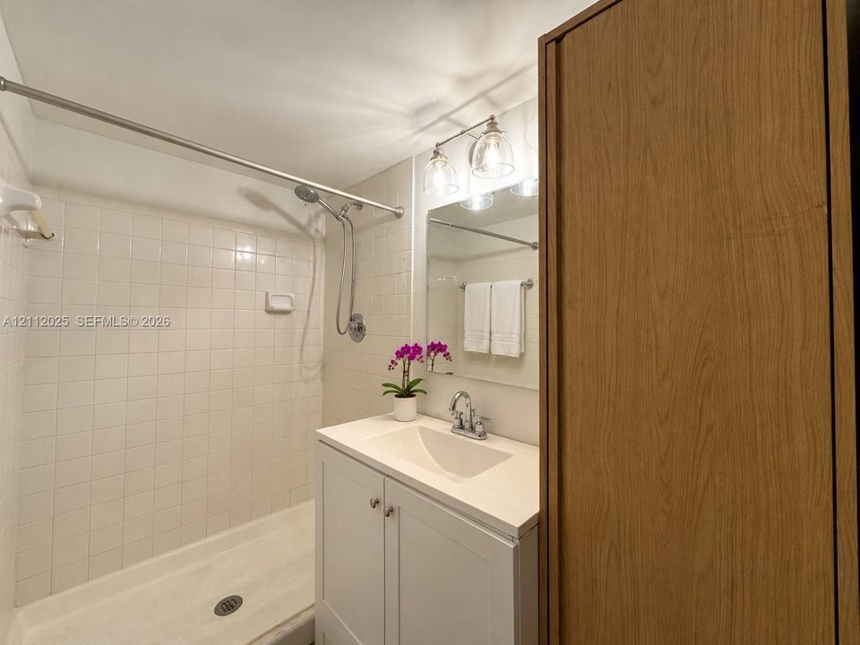 Bathroom featuring a white vanity cabinet with a solid-surface countertop, an integrated sink, a tiled shower surround, and a three-light fixture above a wall-mounted mirror