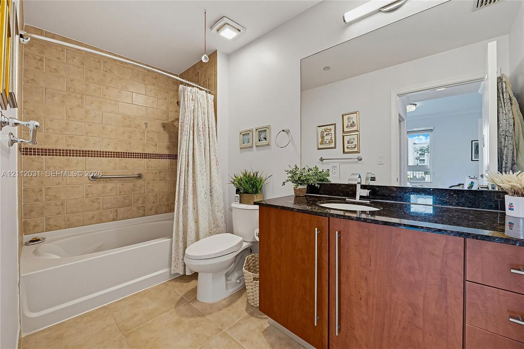 Bathroom featuring a dark stone countertop, wood-finish cabinetry with vertical hardware, an undermount sink, and a tiled shower surround with a built-in grab bar