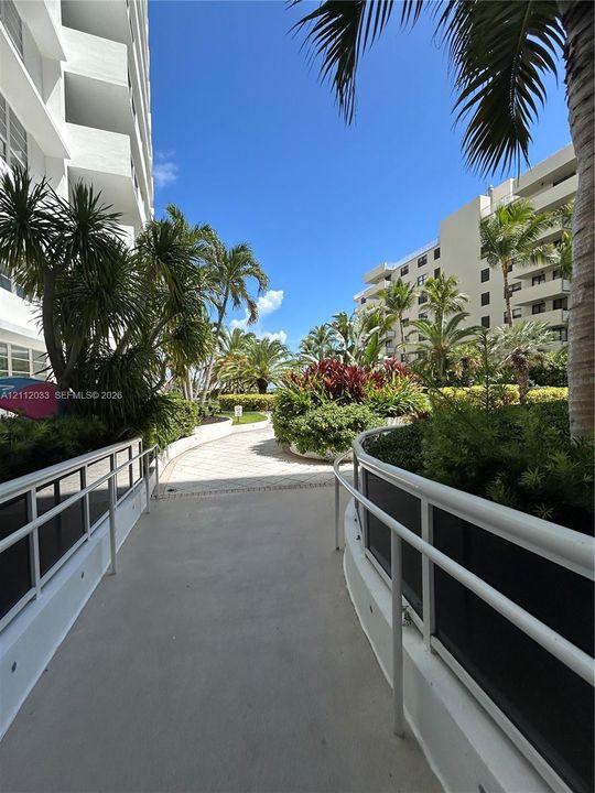 Exterior walkway featuring paved pathways, white metal railings, manicured tropical landscaping, and mature palm trees