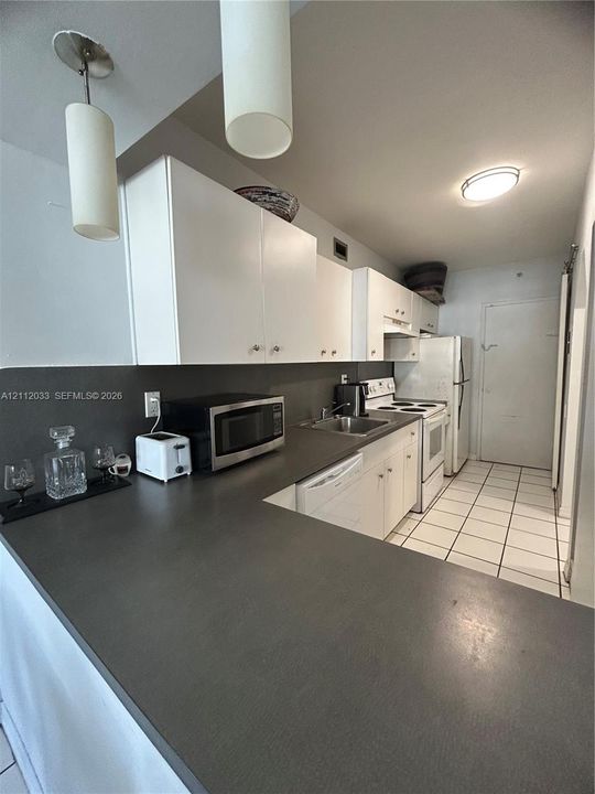 Kitchen featuring dark solid-surface countertops, white cabinetry, integrated stainless steel sink, tiled flooring, and dual cylindrical pendant lighting