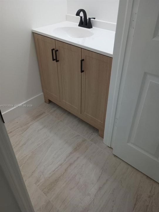 Wood-finish vanity cabinet with matte black hardware, white countertop, integrated basin, matte black faucet, and patterned tile flooring