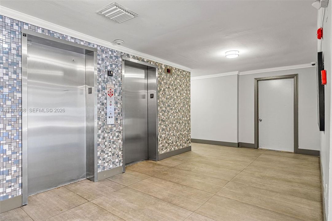 Elevator bank featuring brushed stainless steel doors and mosaic tile wall accents, complemented by large-format tile flooring