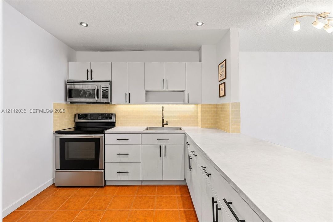 Kitchen featuring white cabinetry with matte black hardware, stainless steel range and microwave, light-toned stone-finish countertops, tile backsplash, and recessed lighting