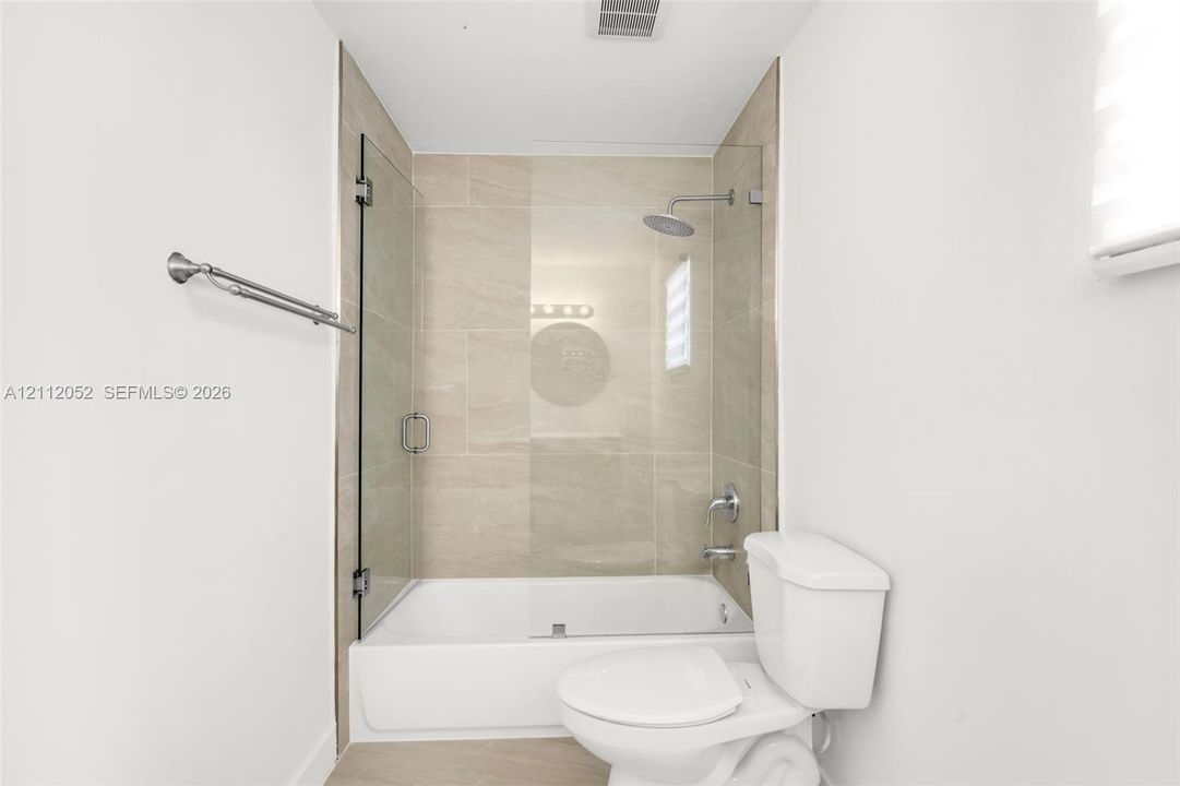 Bathroom featuring a shower-tub combination with a frameless glass door, neutral-tone ceramic wall tiling, a rainfall showerhead, and a wall-mounted metal towel bar