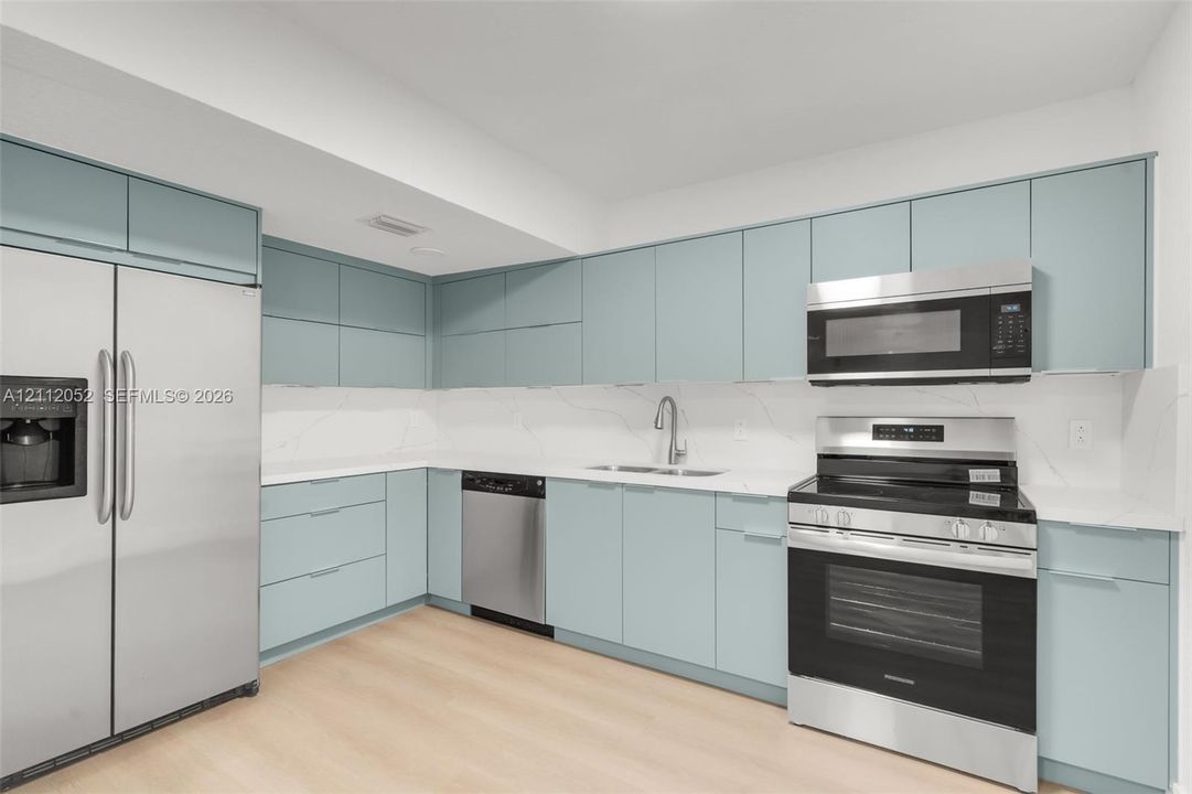 Kitchen featuring matte blue flat-panel cabinetry, light-toned stone-finish countertops, full-height white-veined backsplash, and stainless steel appliances including a built-in microwave and range