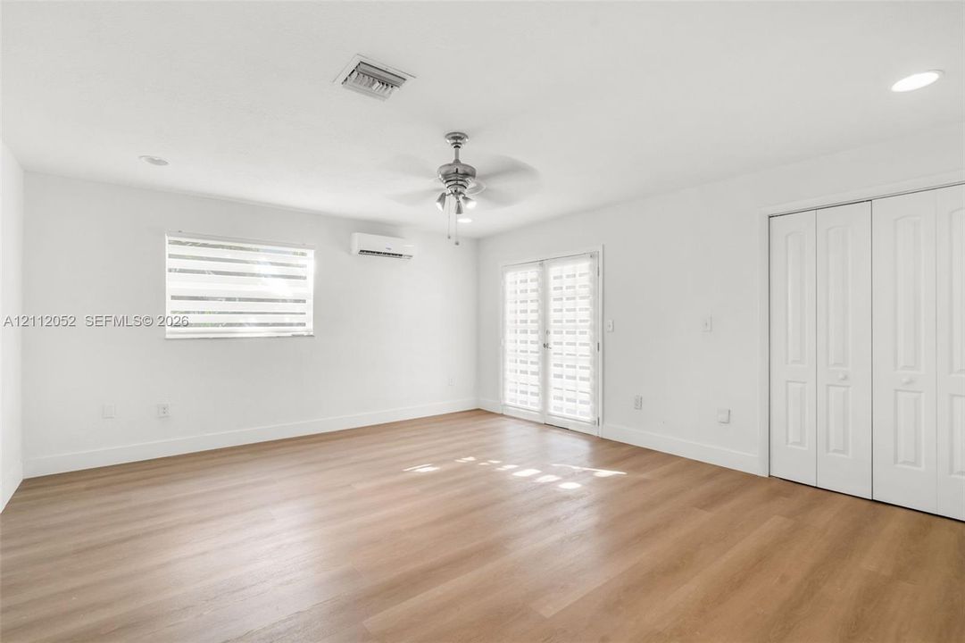 Room featuring wood-finish flooring, a wall-mounted ductless air conditioning unit, a ceiling fan with integrated lighting, French-style doors, and double-panel closet doors