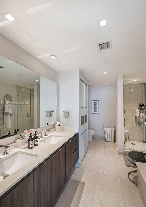 Double vanity featuring wood-finish cabinetry, white countertop with dual integrated sinks, full-width vanity mirror, recessed ceiling lighting, and a glass-enclosed shower