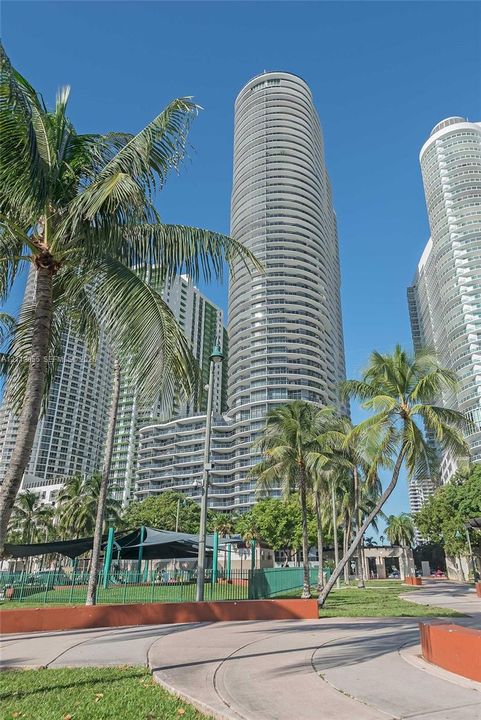 High-rise architectural exterior with cylindrical glass facade, expansive wrap-around balconies, landscaped grounds, palm tree foliage, and paved pedestrian pathways