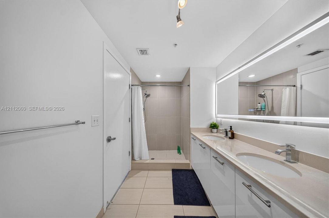 Bathroom featuring a dual-sink vanity with a stone countertop, large wall-to-wall LED-backlit mirror, and a walk-in shower with tile surround