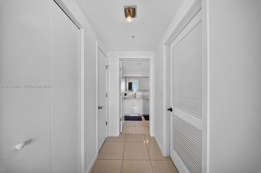Hallway featuring tile flooring, ceiling-mounted light fixture, louvered closet doors, and smooth-finish interior doors with matte black hardware