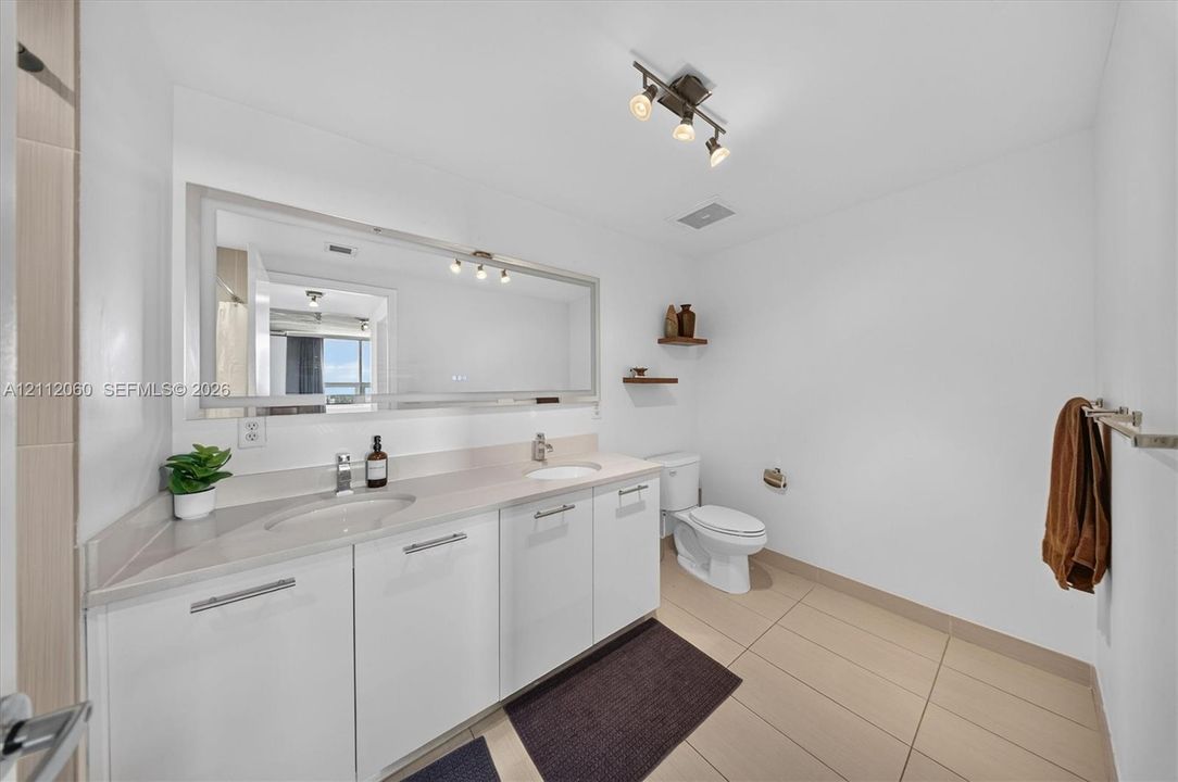 Dual-sink vanity with neutral countertop, expansive wall-mounted mirror, three-light track fixture, and large-format tile flooring