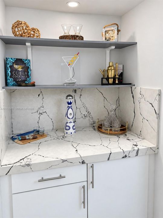 Built-in wet bar featuring stone-patterned surfacing, integrated backsplash, white cabinetry with metallic hardware, and dual grey floating shelves with recessed lighting