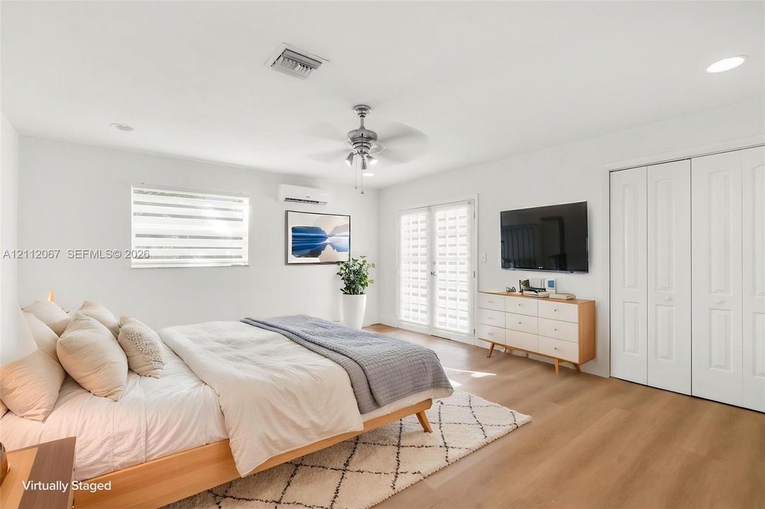 Interior space featuring wood-finish flooring, a wall-mounted ductless climate control unit, a ceiling fan with integrated lighting, dual-panel glass French doors, and double-door closet storage