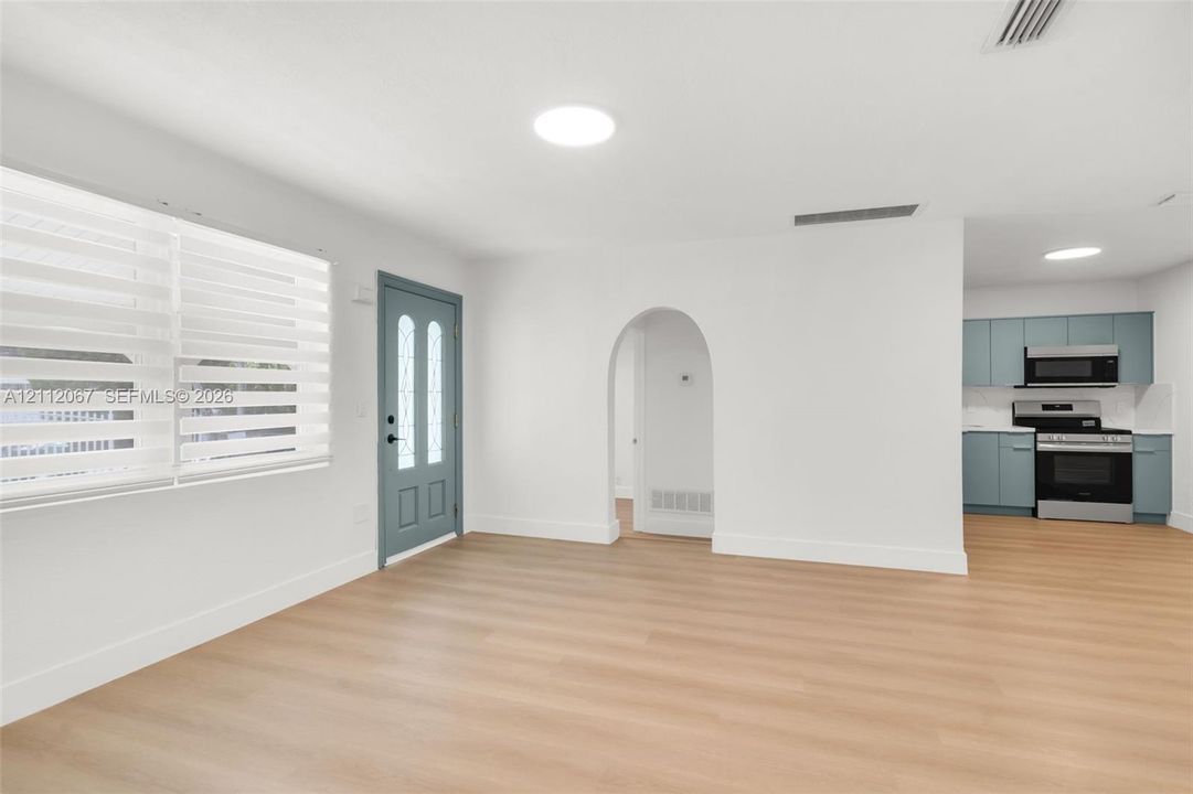 Open-concept interior featuring light wood-finish flooring, an arched entryway, a blue-toned exterior door with glass detailing, and kitchen cabinetry paired with stainless steel appliances